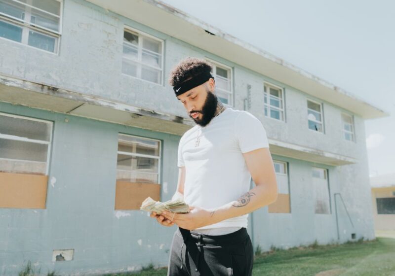 A young man with facial hair counts money in front of a worn building on a sunny day.