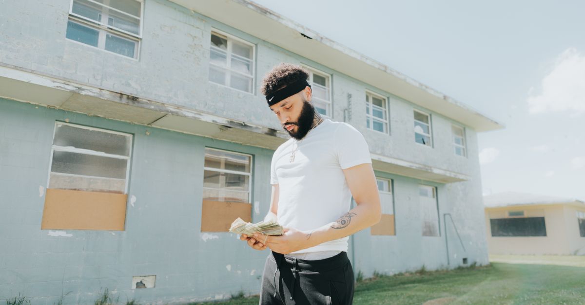 A young man with facial hair counts money in front of a worn building on a sunny day.