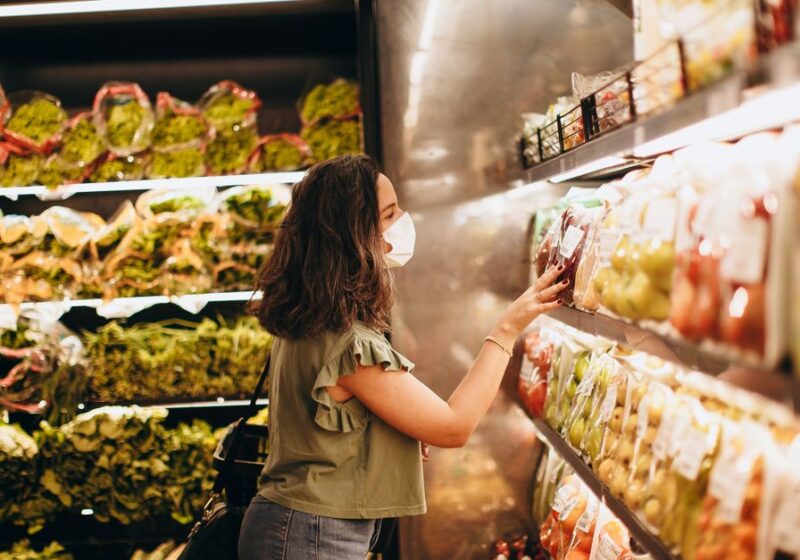 Woman wearing a mask choosing fresh produce in a supermarket aisle.