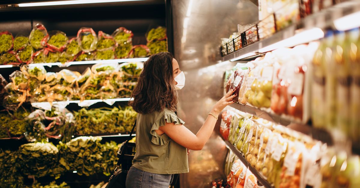 Woman wearing a mask choosing fresh produce in a supermarket aisle.