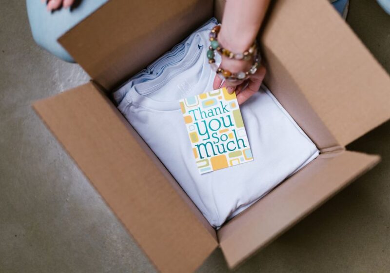 A person packing a white t-shirt with a thank you card into a cardboard box.