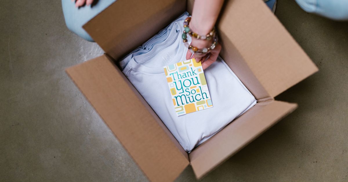 A person packing a white t-shirt with a thank you card into a cardboard box.