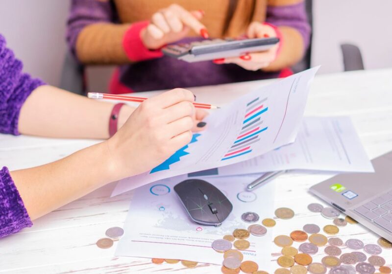 Two women analyzing financial charts with a laptop and coins on desk in an office setting.