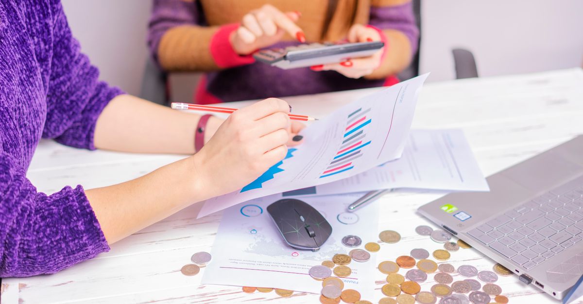 Two women analyzing financial charts with a laptop and coins on desk in an office setting.