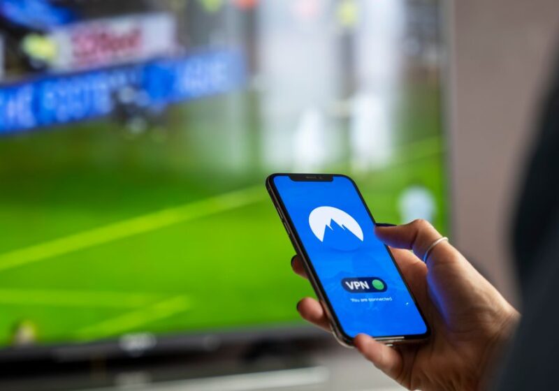 Close-up of a person holding a smartphone with a VPN app, streaming sports on TV.