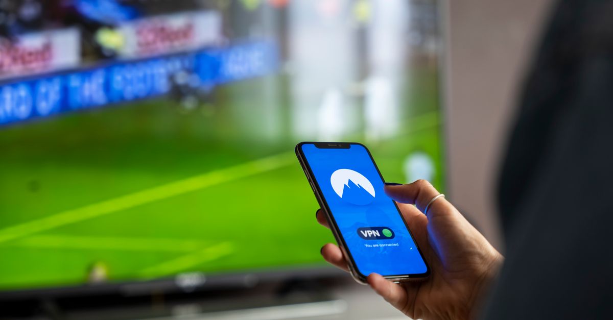 Close-up of a person holding a smartphone with a VPN app, streaming sports on TV.