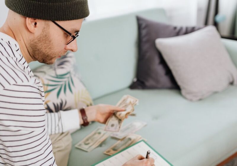 Adult man with beanie counting cash and making notes on a couch.