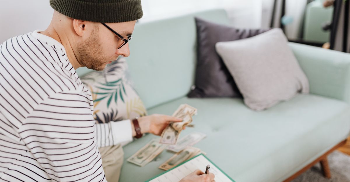 Adult man with beanie counting cash and making notes on a couch.