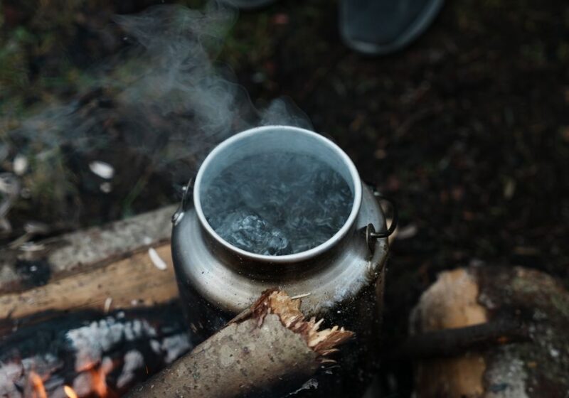 A silver kettle steaming as it boils water over a rustic campfire in the woods. Outdoor camping scene.