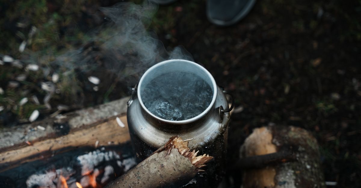 A silver kettle steaming as it boils water over a rustic campfire in the woods. Outdoor camping scene.