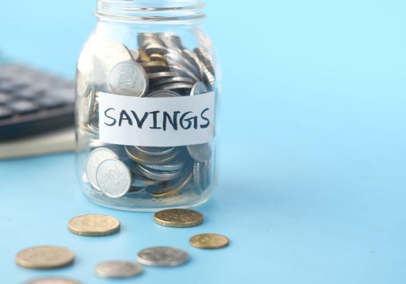 Glass jar labeled 'Savings' filled with coins, beside a calculator on a blue background.