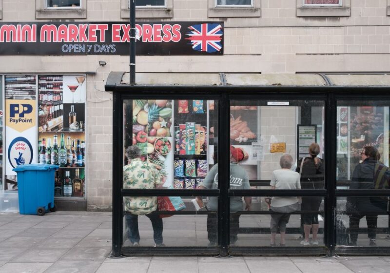 A bustling city bus stop in front of a mini market with people waiting and street advertisements.