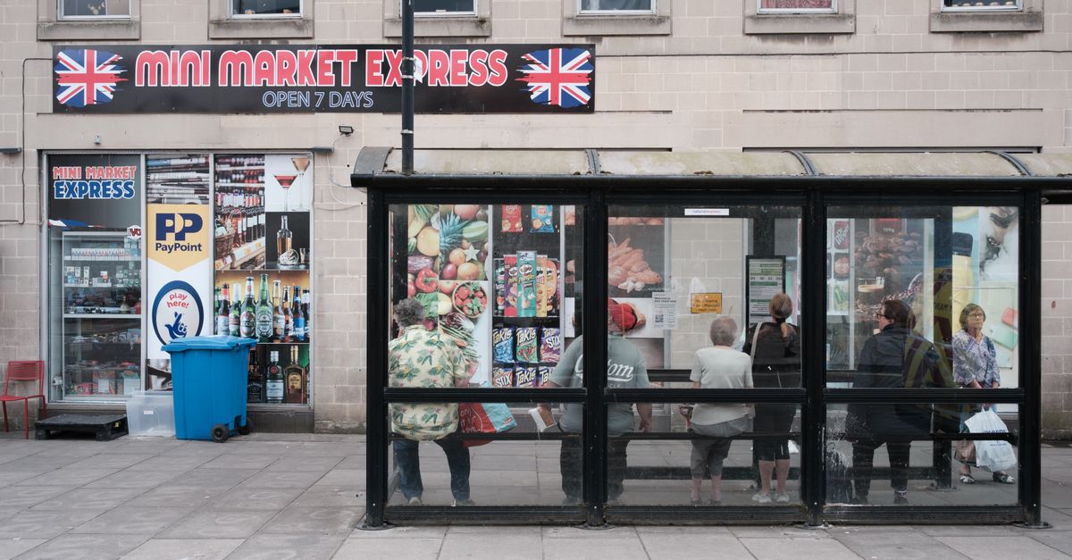 A bustling city bus stop in front of a mini market with people waiting and street advertisements.