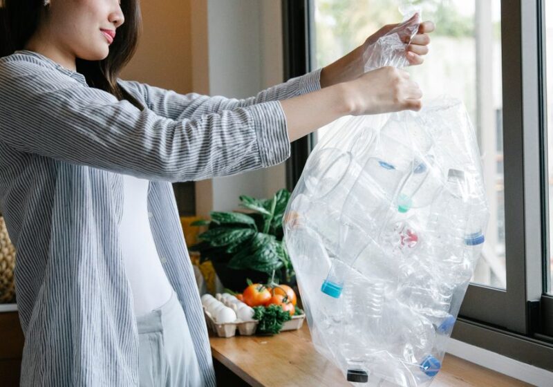 Side view of positive Asian female with transparent bag full of plastic bottles for recycling standing in light kitchen at home