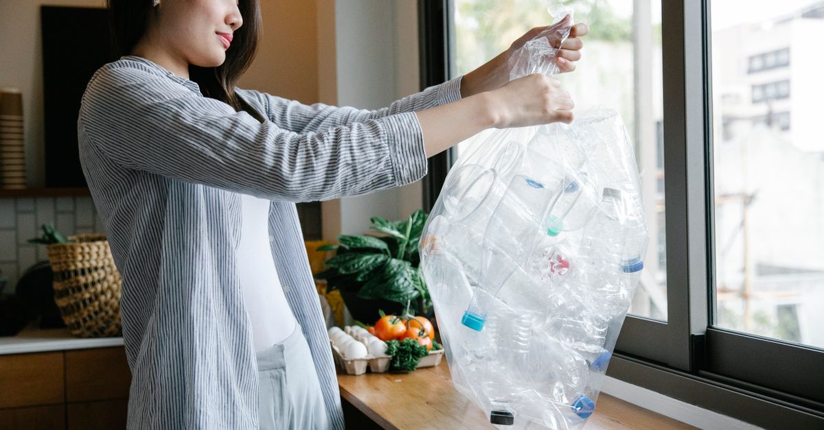 Side view of positive Asian female with transparent bag full of plastic bottles for recycling standing in light kitchen at home